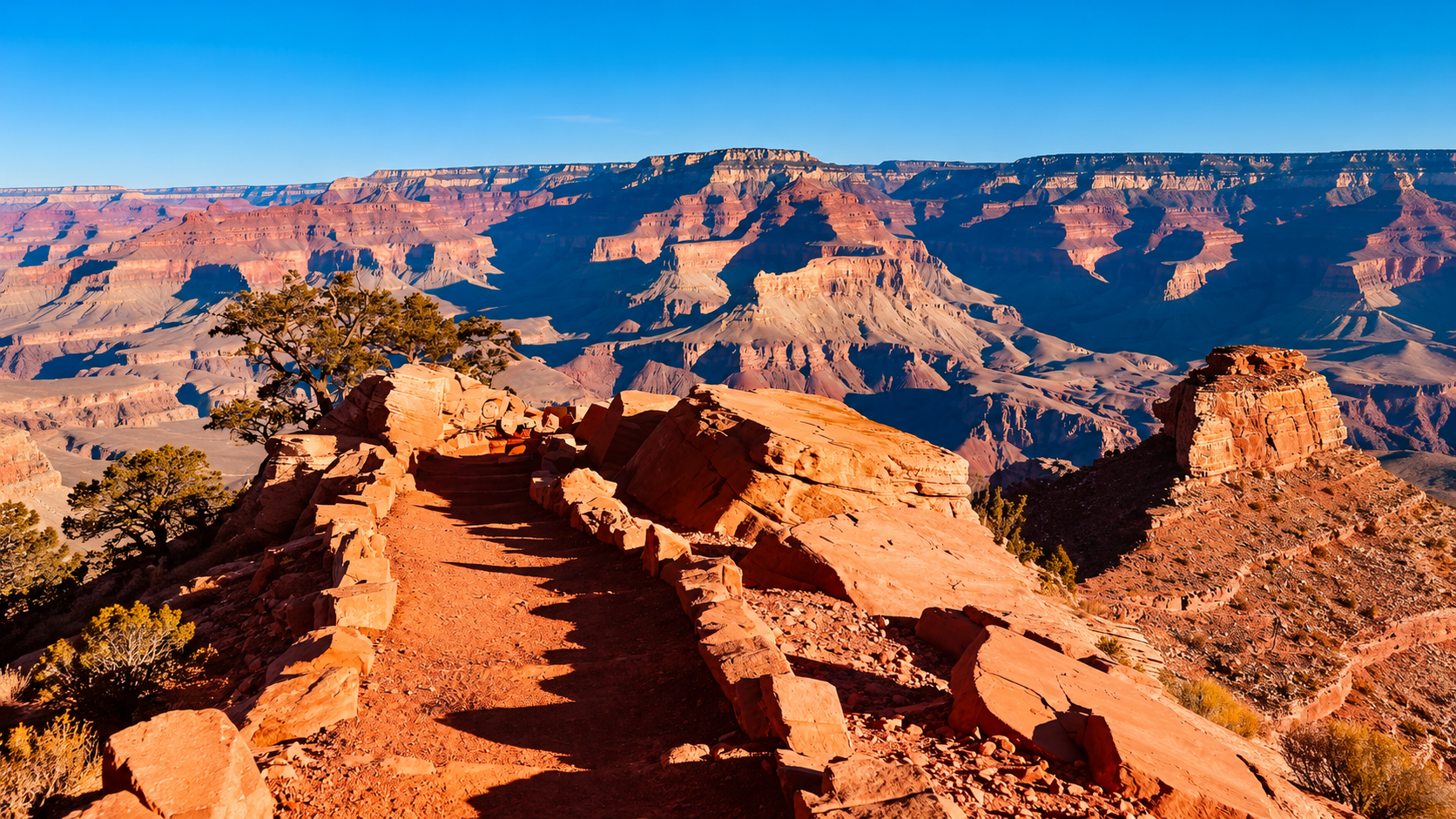 A red-rock trail on South Kaibab winding toward a sweeping view of the Grand Canyon under a clear blue sky